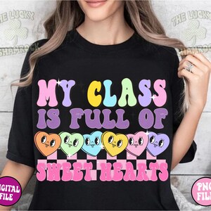 May include: Black t-shirt with the text "My Class Is Full Of Sweethearts" in rainbow colors. The text is surrounded by a row of six cartoon candy hearts with faces.