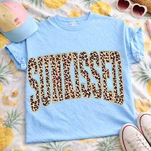 May include: A light blue t-shirt with the word "SUNKISSED" in large, leopard print letters. The shirt is folded, with a baseball cap and sunglasses in the background. The shirt is on a pineapple-patterned surface.