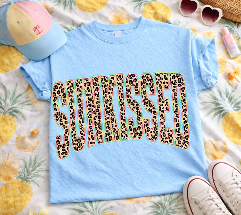 May include: A light blue t-shirt with the word "SUNKISSED" in large, leopard print letters. The shirt is folded, with a baseball cap and sunglasses in the background. The shirt is on a pineapple-patterned surface.