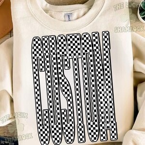 May include: A cream-colored crewneck sweatshirt featuring the word CUSTOM printed in a large, bold font. The letters are filled with a black and white checkered pattern. The garment has a ribbed neckline and a Gildan brand label at the collar.