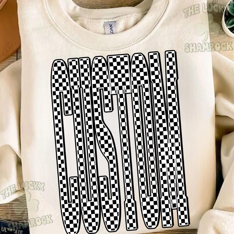 May include: A cream-colored crewneck sweatshirt featuring the word CUSTOM printed in a large, bold font. The letters are filled with a black and white checkered pattern. The garment has a ribbed neckline and a Gildan brand label at the collar.