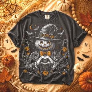 May include: A charcoal gray t-shirt featuring a graphic illustration of a pumpkin-headed figure wearing a witch hat and a suit with a bow tie. The figure is making a heart shape with its hands, surrounded by bats, stars, and autumn leaves.