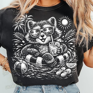 May include: Black graphic t-shirt featuring a raccoon wearing sunglasses and floating in a pool with a drink. The raccoon is in a black and white inner tube. Palm trees, a beach ball, and flip-flops are also in the design.