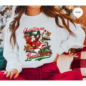 May include: White sweatshirt with a Christmas themed design featuring Santa Claus riding in a sleigh with a pelican and alligator. The design includes the text "Christmas Louisiana".