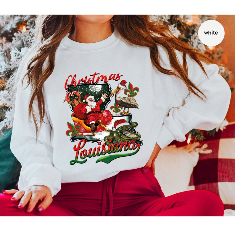 May include: White sweatshirt with a Christmas themed design featuring Santa Claus riding in a sleigh with a pelican and alligator. The design includes the text "Christmas Louisiana".