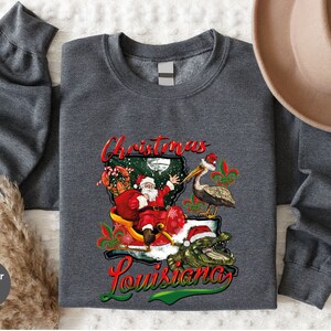 May include: Heather dark grey sweatshirt with a Christmas in Louisiana design. Santa Claus is riding in a sleigh with a pelican and alligator. The design includes the text "Christmas Louisiana".