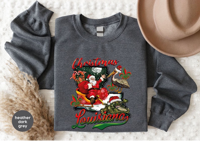 May include: Heather dark grey sweatshirt with a Christmas in Louisiana design. Santa Claus is riding in a sleigh with a pelican and alligator. The design includes the text "Christmas Louisiana".