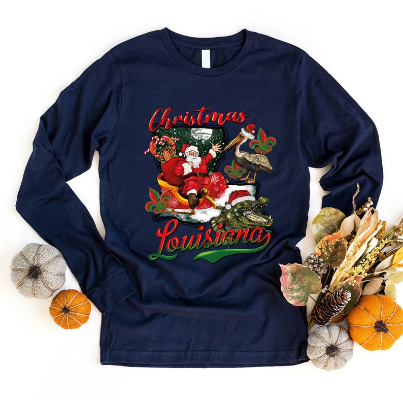May include: A navy blue long-sleeved t-shirt with a Christmas themed design featuring Santa Claus in a sleigh with a pelican and alligator. The design also includes the text "Christmas Louisiana".