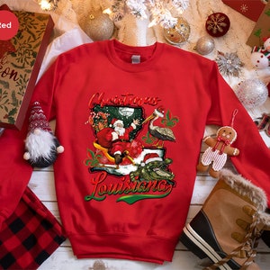 May include: Red Christmas sweatshirt with a graphic of Santa Claus riding a sleigh in the shape of Louisiana, with a pelican and alligator. The text "Christmas Louisiana" is printed on the sweatshirt.