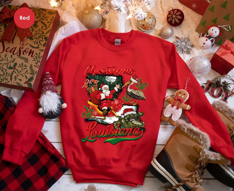 May include: Red Christmas sweatshirt with a graphic of Santa Claus riding a sleigh in the shape of Louisiana, with a pelican and alligator. The text "Christmas Louisiana" is printed on the sweatshirt.