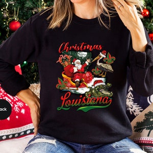May include: Black sweatshirt with a Christmas themed design featuring Santa Claus riding in a sleigh with an alligator, a pelican, and the text "Christmas Louisiana".