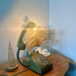 May include: A green rotary phone with a white dial and a light on the receiver. The phone is on a wooden table with a small gold wire tree ornament next to it.