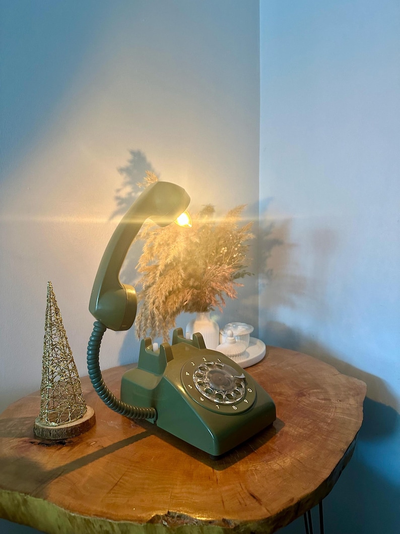 May include: A green rotary phone with a white dial and a light on the receiver. The phone is on a wooden table with a small gold wire tree ornament next to it.