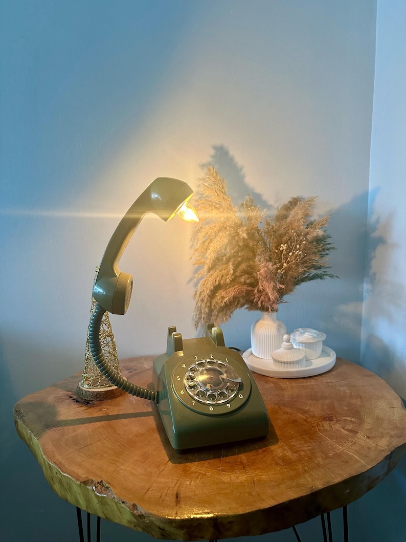 May include: A green rotary phone with a white dial and a light bulb in the receiver. The phone is on a wooden table with a vase of dried flowers and a white ceramic object.