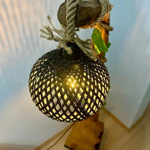 Op de afbeelding: Een geweven zwarte lampenkap met een gloeilamp erin, hangend aan een houten tak met touw en groene bladeren.