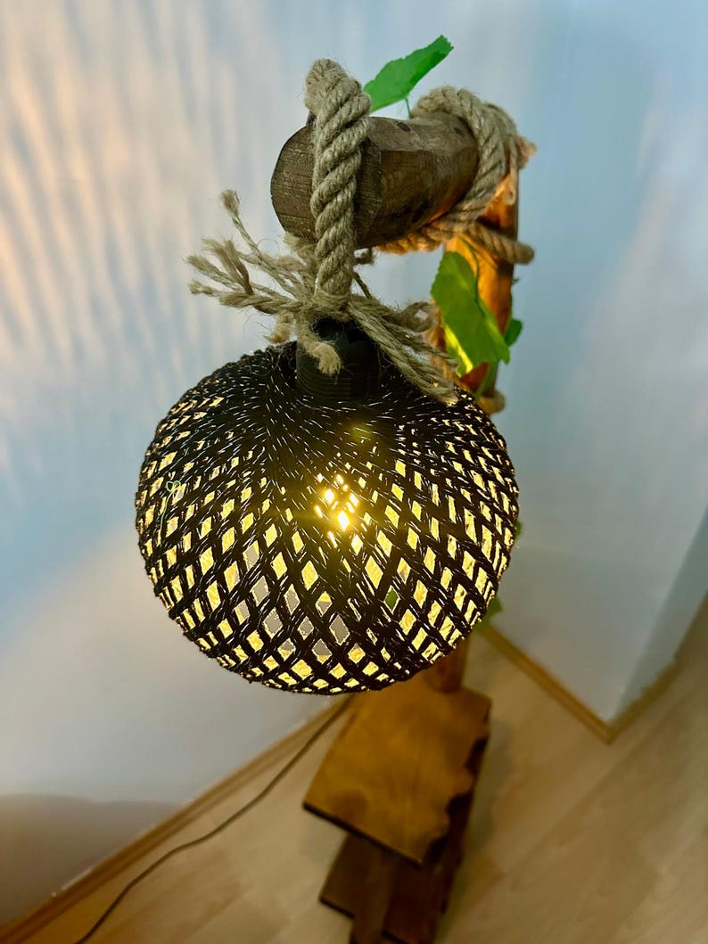Op de afbeelding: Een geweven zwarte lampenkap met een gloeilamp erin, hangend aan een houten tak met touw en groene bladeren.