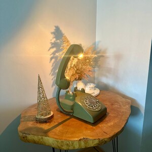 May include: A green rotary phone lamp on a wooden table with a gold decorative piece. The phone has a white dial and a light bulb in the receiver.