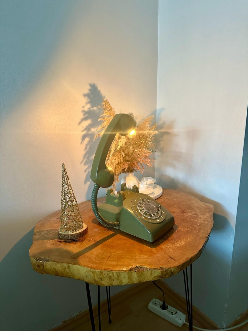 May include: A green rotary phone lamp on a wooden table with a gold decorative piece. The phone has a white dial and a light bulb in the receiver.
