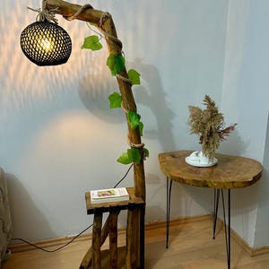 Op de afbeelding: Een houten vloerlamp met een zwarte geweven lampenkap en een touw om de basis gewikkeld. De lamp is versierd met kunstmatige groene bladeren en heeft een klein houten plankje. De lamp staat naast een kleine houten tafel met zwarte metalen poten.