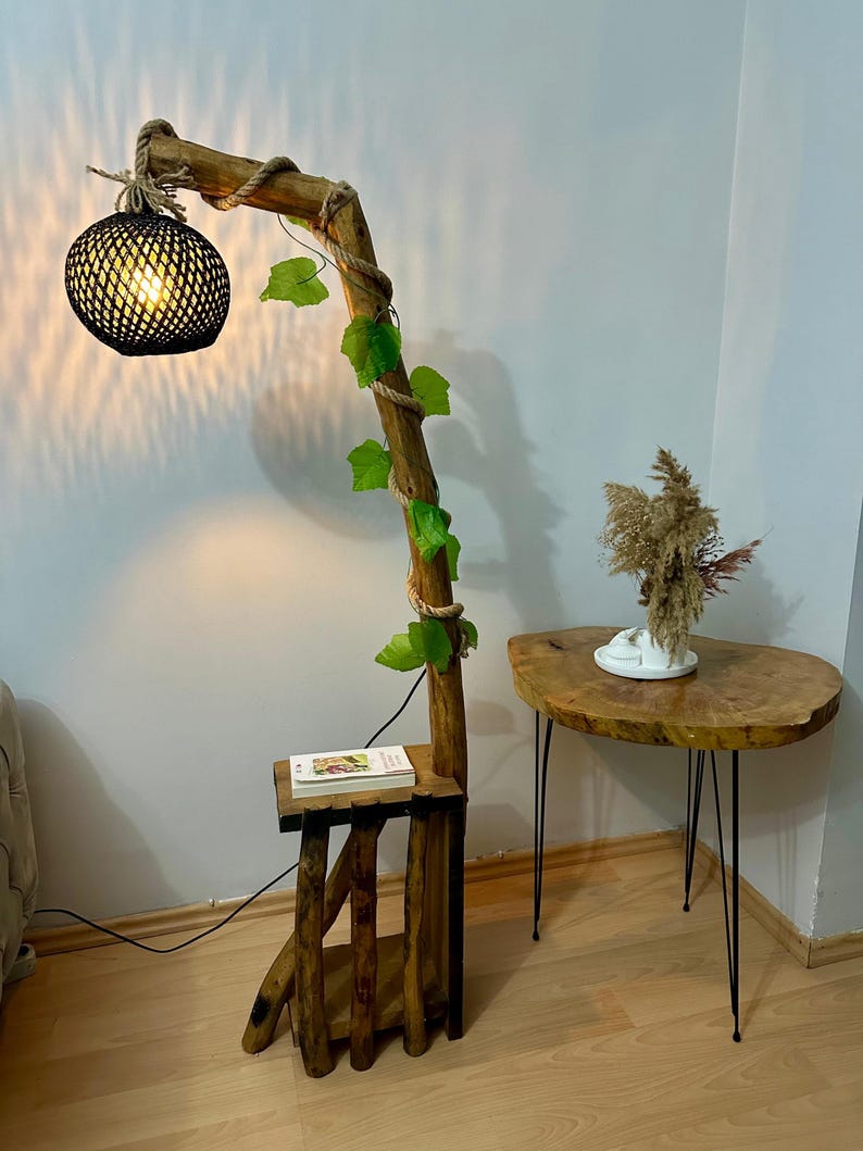 Op de afbeelding: Een houten vloerlamp met een zwarte geweven lampenkap en een touw om de basis gewikkeld. De lamp is versierd met kunstmatige groene bladeren en heeft een klein houten plankje. De lamp staat naast een kleine houten tafel met zwarte metalen poten.