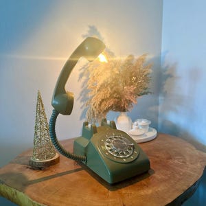 May include: A green rotary phone with a light bulb in the receiver. The phone is on a wooden table with a small golden tree and dried flowers in a vase.