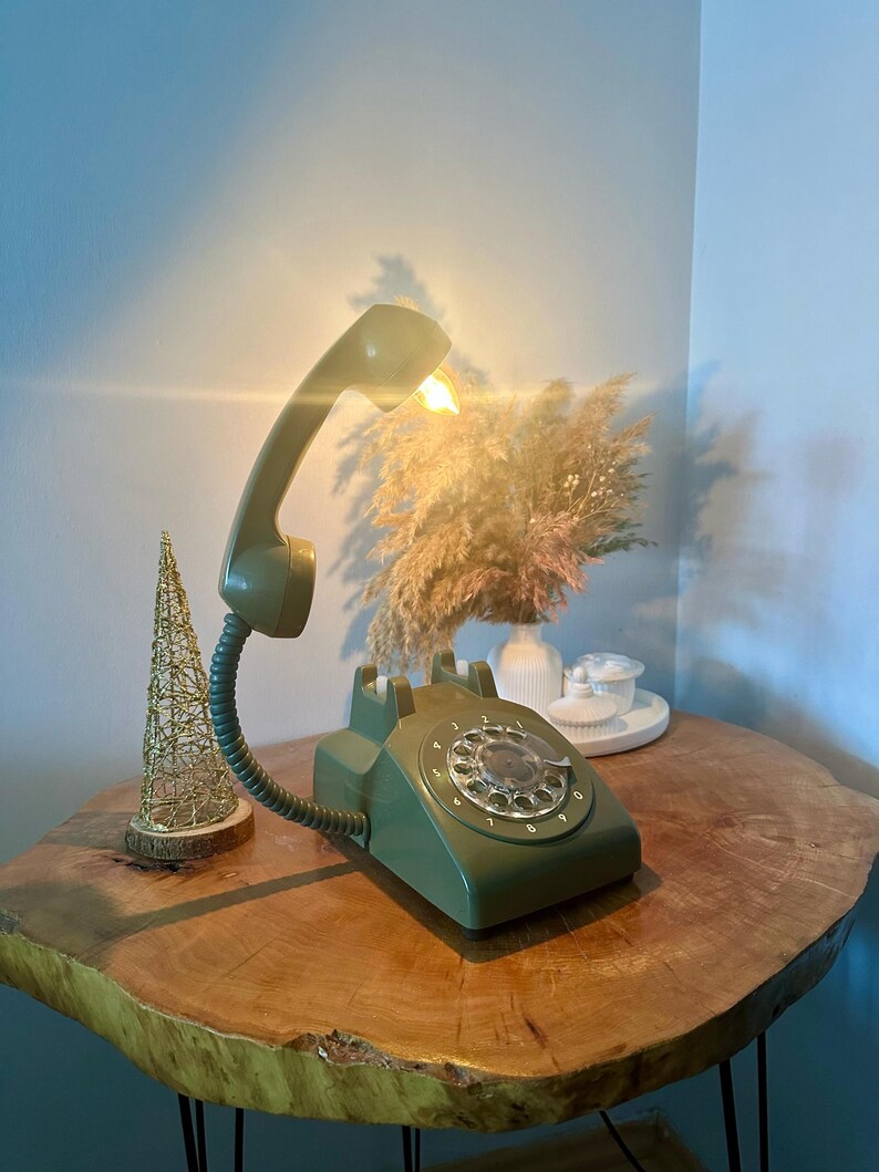 May include: A green rotary phone with a light bulb in the receiver. The phone is on a wooden table with a small golden tree and dried flowers in a vase.