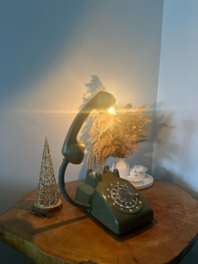 May include: A green rotary phone with a flexible arm and a light bulb on the receiver. The phone is on a wooden surface with a small gold wire tree and a vase of dried flowers.