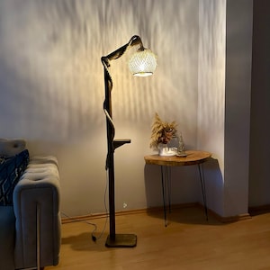 May include: A wooden floor lamp with a rope-like stem and a woven shade. The lamp is turned on and casting a warm glow on the surrounding area. The lamp is next to a small wooden side table with a black metal frame.
