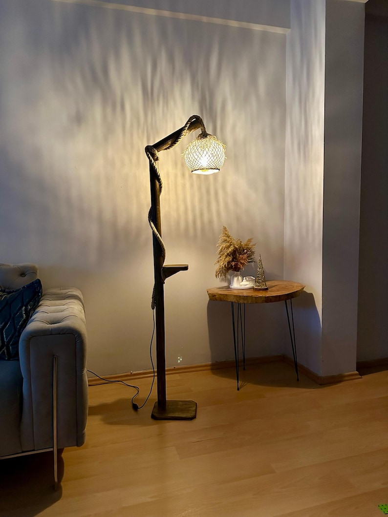 May include: A wooden floor lamp with a rope-like stem and a woven shade. The lamp is turned on and casting a warm glow on the surrounding area. The lamp is next to a small wooden side table with a black metal frame.