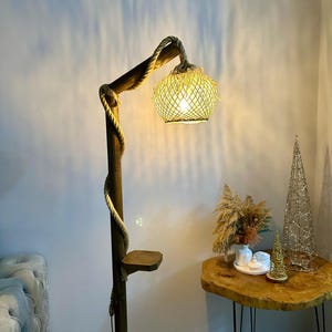May include: A wooden floor lamp with a rope wrapped around the pole and a woven shade. The lamp is lit and casting a warm glow. The lamp has a small shelf on the side.