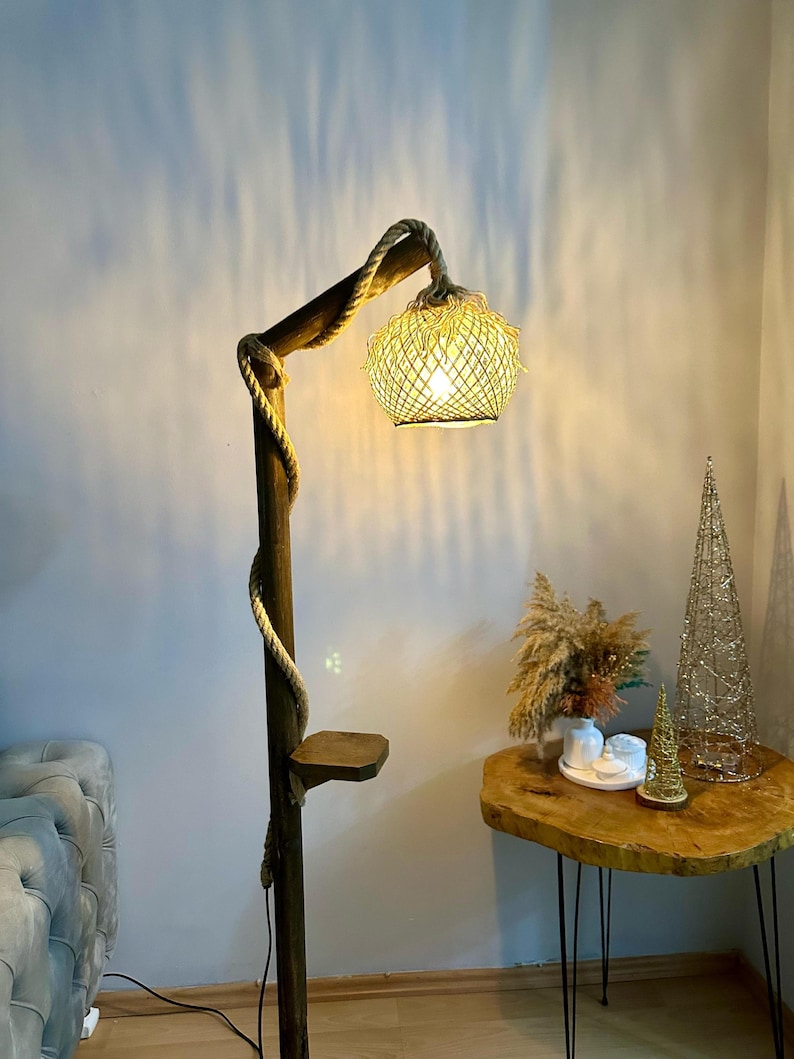 May include: A wooden floor lamp with a rope wrapped around the pole and a woven shade. The lamp is lit and casting a warm glow. The lamp has a small shelf on the side.