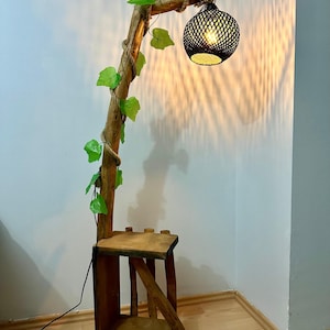 Op de afbeelding: Een houten vloerlamp met een natuurlijk takontwerp en een zwarte geweven lampenkap. De lamp heeft een plank en is versierd met groene bladeren.