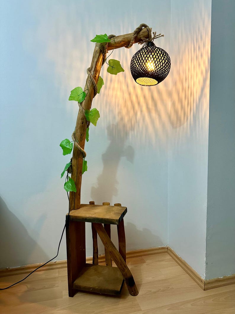 Op de afbeelding: Een houten vloerlamp met een natuurlijk takontwerp en een zwarte geweven lampenkap. De lamp heeft een plank en is versierd met groene bladeren.