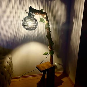 Op de afbeelding: Een houten vloerlamp met een geweven lampenkap en een tak die met touw is omwikkeld. De lamp is aan en werpt een warme gloed op de muur. De lamp staat op een houten voet met een klein plankje.