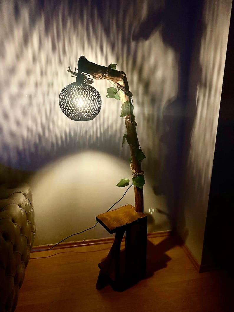 Op de afbeelding: Een houten vloerlamp met een geweven lampenkap en een tak die met touw is omwikkeld. De lamp is aan en werpt een warme gloed op de muur. De lamp staat op een houten voet met een klein plankje.