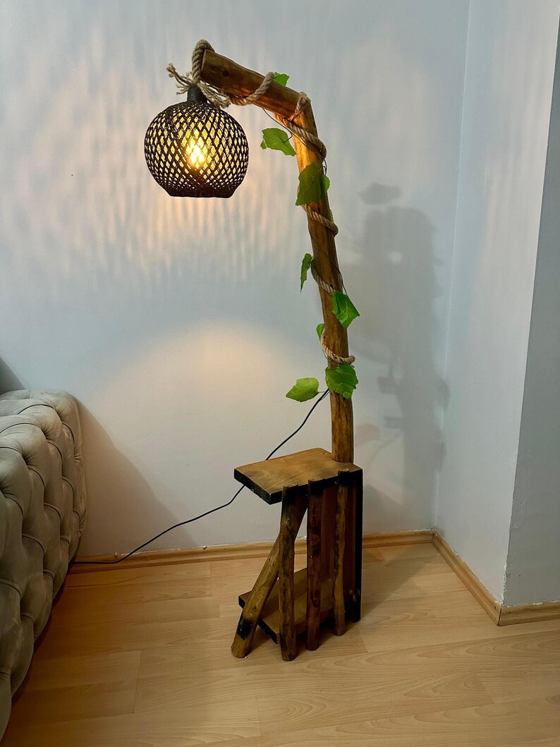 Op de afbeelding: Een houten vloerlamp met een natuurlijke houten basis en een geweven lampenkap. De lamp is gevormd als een tak met touw eromheen gewikkeld en kunstmatige bladeren. De lamp heeft een klein houten plankje bevestigd aan de basis.