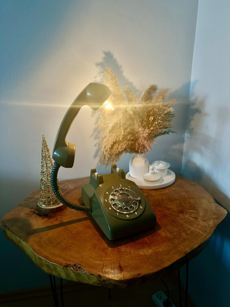 May include: A green rotary phone with a white dial and a green handset. The phone is on a wooden table with a dried flower arrangement in the background.