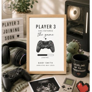 Può includere: Una stampa incorniciata con il testo PLAYER 3 HAS ENTERED the game, un'illustrazione di un controller per videogiochi nero, una barra di caricamento e il testo BABY SMITH ARRIVING MAY 2025. Il design è presentato in una cornice in legno chiaro.