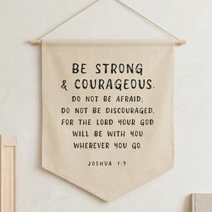 Puede incluir: Una escena de decoración de pared de guardería con una cuna de madera, una pancarta con el texto "BE STRONG & COURAGEOUS" y una estantería con un libro y juguetes. La pancarta es de color crema con texto negro.