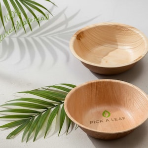 May include: Two wooden bowls with a natural wood grain pattern. One bowl has the text "PICK A LEAF" and a leaf graphic. The image also includes green palm leaves and the text "100% Natural Material!"