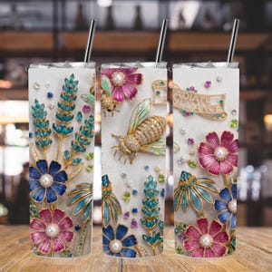 Puede incluir: Tres vasos de acero inoxidable con diseños florales y de abejas. Los diseños presentan adornos azules, rosas y dorados con piedras preciosas y perlas. Cada vaso tiene una pajita plateada y el texto "RADIATE ELEGANCE".