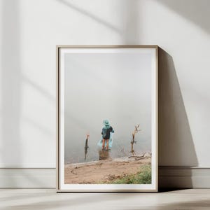 May include: Framed art print featuring a person standing in a body of water, wearing a hat and holding a blue inflatable. The artwork is displayed against a white wall, with natural light and shadows. The frame is a neutral color.