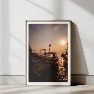 May include: Framed print of boats on the water at sunset. The sun is bright and the water reflects the light. The boats are wooden and have a flag on top. The print is in a neutral-toned frame.