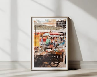 May include: A street food vendor in a bustling market setting, preparing food on a cart with a red umbrella overhead. The vendor is wearing a blue shirt and a red apron, and there are other people in the background.