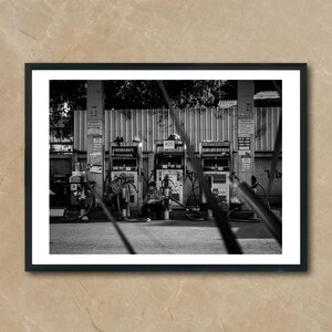 May include: A black and white photo of three gas pumps at a gas station. The pumps are in front of a brick wall and there are trees in the background.