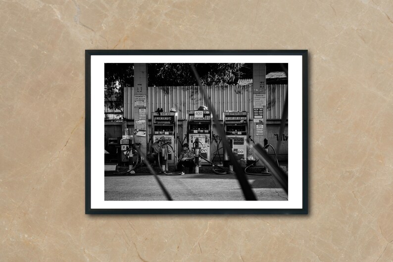 May include: A black and white photo of three gas pumps at a gas station. The pumps are in front of a brick wall and there are trees in the background.