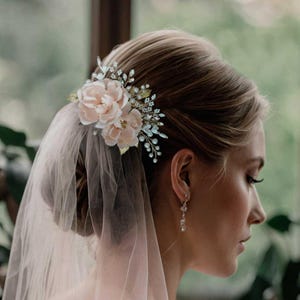 May include: A bridal hair accessory with blush fabric flowers, pearls, and silver details. The floral arrangement is attached to the hair with a silver comb. A sheer veil is draped over the hair and shoulders.