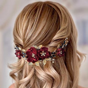 May include: A burgundy floral hair comb with gold leaves and dark blue accents. The comb features three large burgundy flowers with gold centers and is set against wavy blonde hair. A decorative hair accessory.