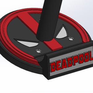 May include: A black and red Deadpool-themed stand. The stand features the Deadpool logo and the word "DEADPOOL" in red letters. The stand is designed with a circular base and a rectangular front.