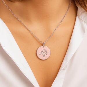 Pu&ograve; includere: Una collana d'argento con un ciondolo circolare con una silhouette nera di testa di cavallo su sfondo rosa. La collana &egrave; indossata su una camicia bianca.
